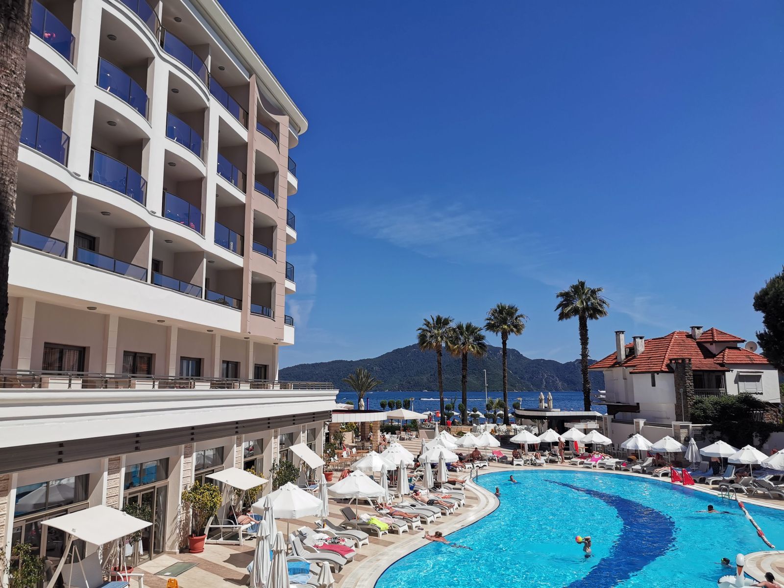 A hotel pool by the sea, sunbeds and parasols full of people
